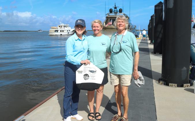 From left: Cathy Chapman, Fernandina Beach Marina Manager, Debra Strobel and Candis Whitney, directors of Amelia Island Whale Ambassadors, pose Nov. 8 at the marina with one of the totes that are being passed out to boaters at the marina as a part of a new inititive. The tote encourages boaters to take precautions, slow down and keep a look out for whales. Photo by Ashley Chandler/News-Leader