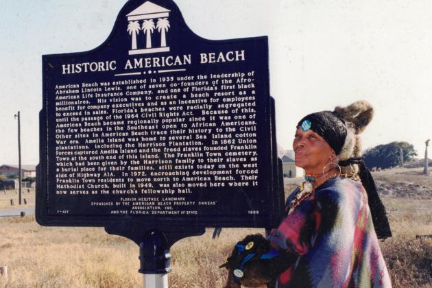 MaVynee Oshun Betsch, "The Beach Lady" stands beside an American Beach historical marker. Photo from A.L. Lewis Museum at American Beach