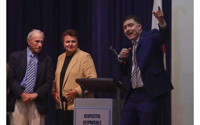 Howard Pines, left, and Dr. Kathy Burns look on as moderator of the event, Bradley Bean announces the winning team of the 2025 Constitution Bowl. Photo by Ashley Chandler/News-Leader