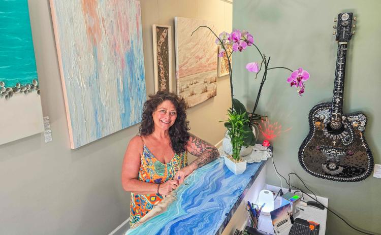 Julie Delfs stands beside the custom-made front desk at Art Amelia, her artwork hangs on the wall to each side of her. Photo by Ashley Chandler/News-Leader